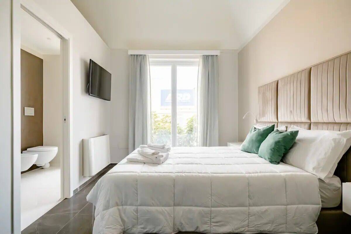 A modern bedroom with a large bed, a flat-screen TV, a window with curtains, and a view of the outdoors.
