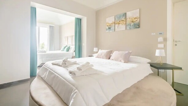 A modern hotel room with a large bed, a bedside table with a lamp, and a view of the adjacent room. - SHoruq (Marsala)