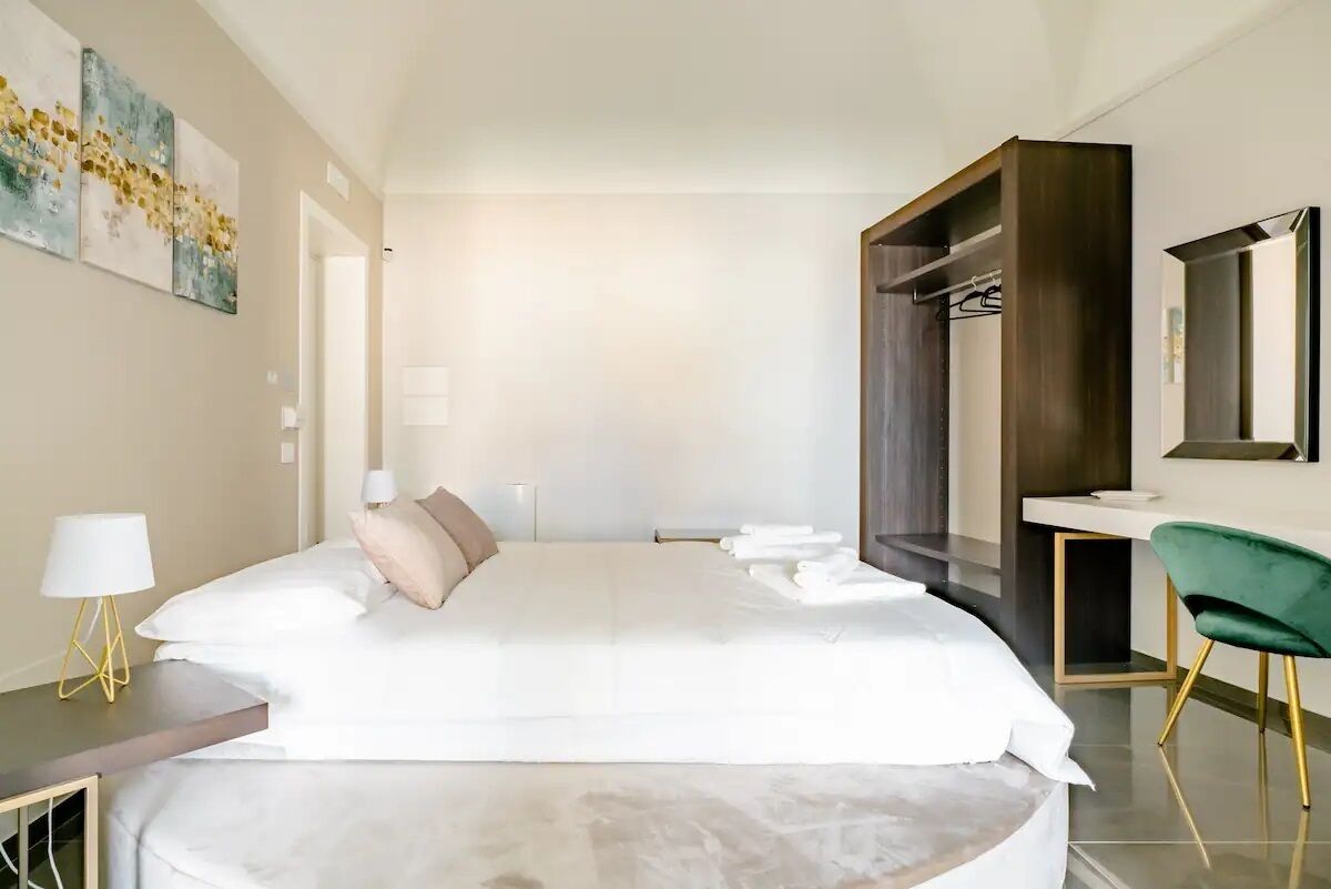 Une chambre d’hôtel moderne avec un grand lit, une table de chevet avec une lampe, un fauteuil vert, une armoire et un miroir au mur.