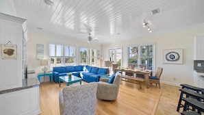 A spacious living room with a blue sectional sofa, a coffee table, and patterned armchairs. There is a dining area with a wooden table and chairs. - "Surfer Dog" | Beachfront Pool | Includes Beach Chairs & Bikes (Inlet Beach)