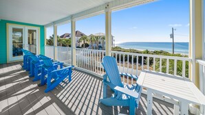 A covered outdoor area with blue Adirondack chairs and a table, overlooking a beach and ocean. - "Surfer Dog" | Beachfront Pool | Includes Beach Chairs & Bikes (Inlet Beach)