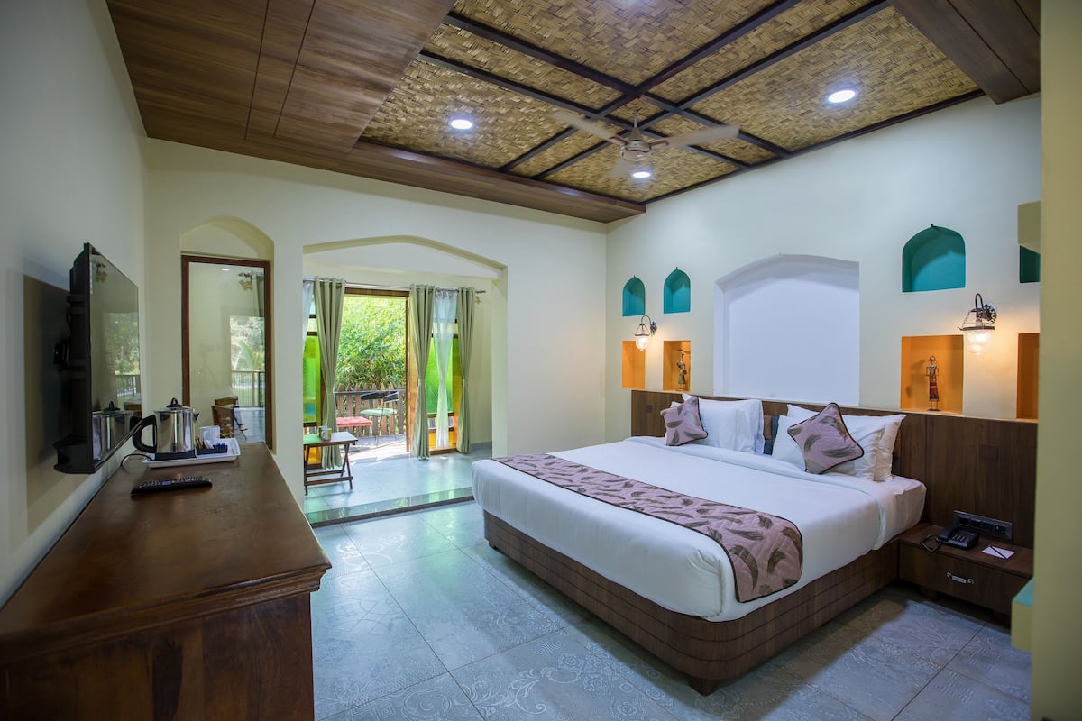 A spacious bedroom with a large bed, a wooden dresser, a television, and a balcony with a view of greenery.