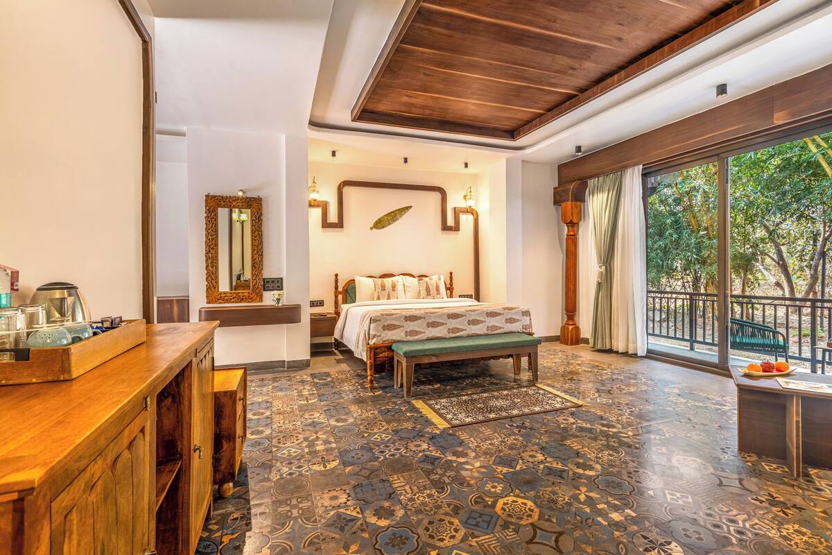 A bedroom with a bed, bench, wooden desk, and a large window with a view of trees.