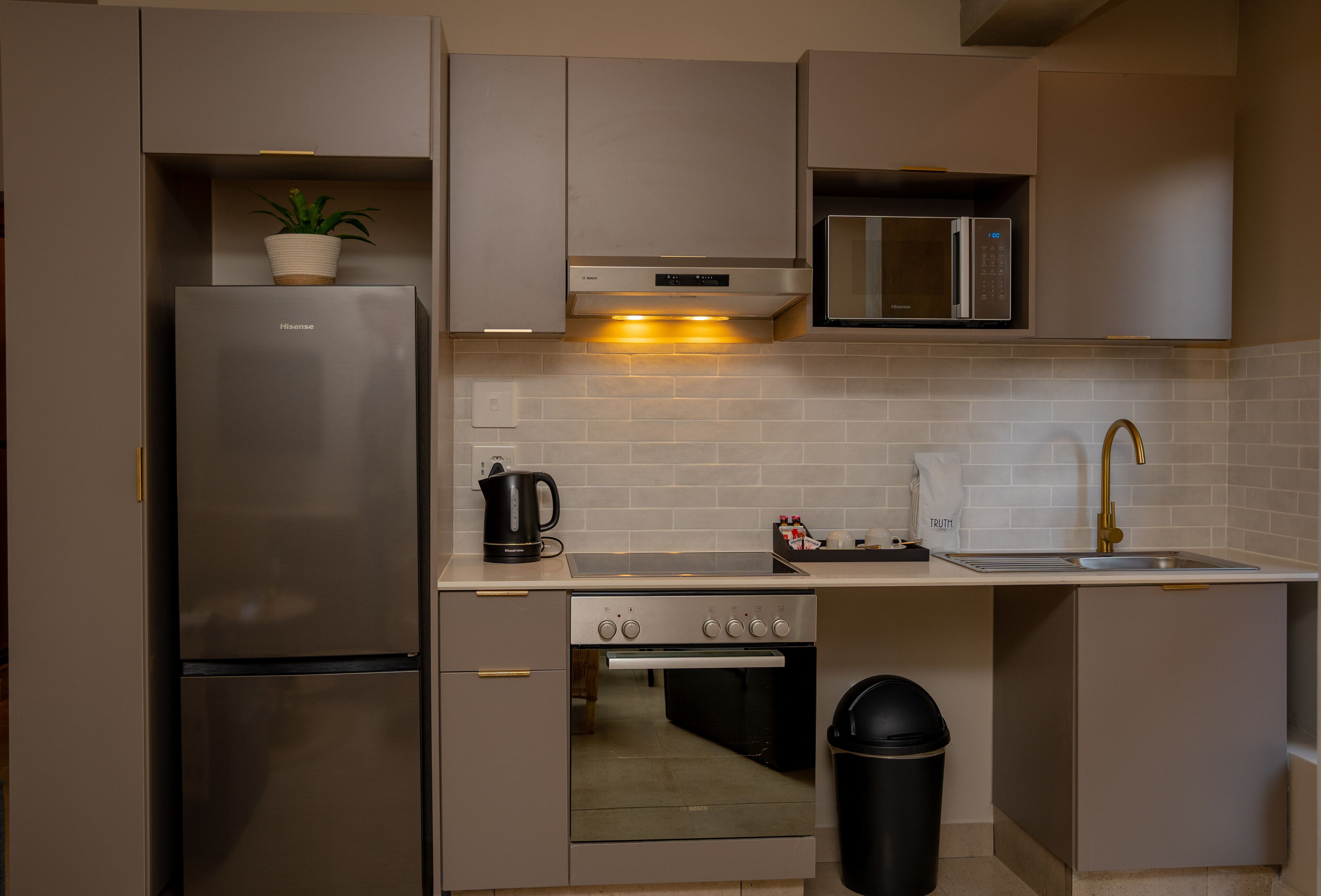 A compact kitchen with a stainless steel refrigerator, a microwave, an oven, and a sink with a gold faucet.