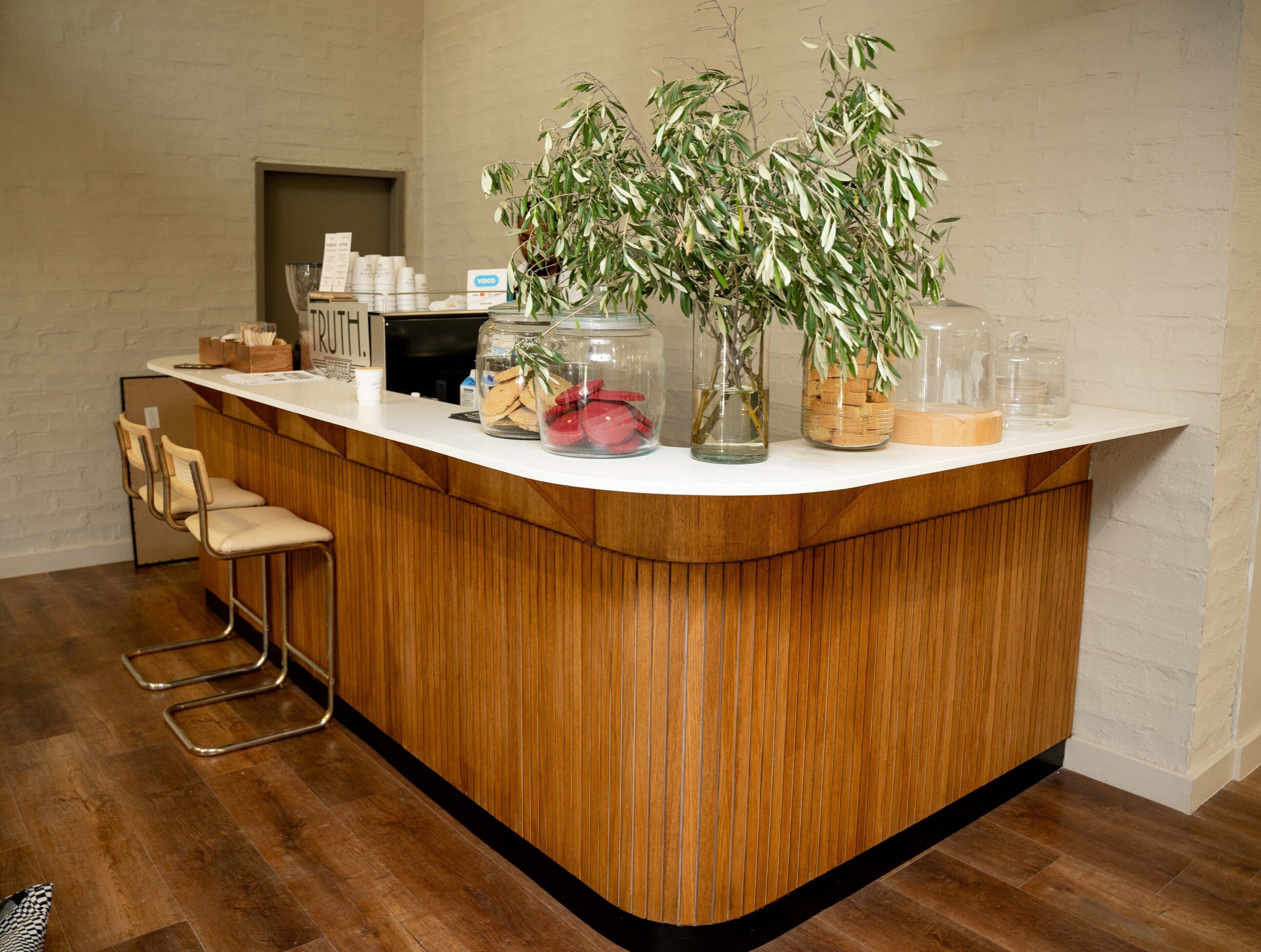 A kitchen counter with a wooden finish, a coffee machine, and a sign that reads "RUTH." There are also two chairs and a potted plant on the counter.