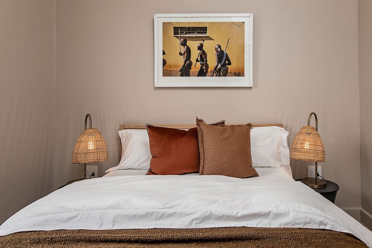 Een netjes opgemaakt bed met witte lakens en twee bruine kussens, met aan weerszijden twee rieten lampen.