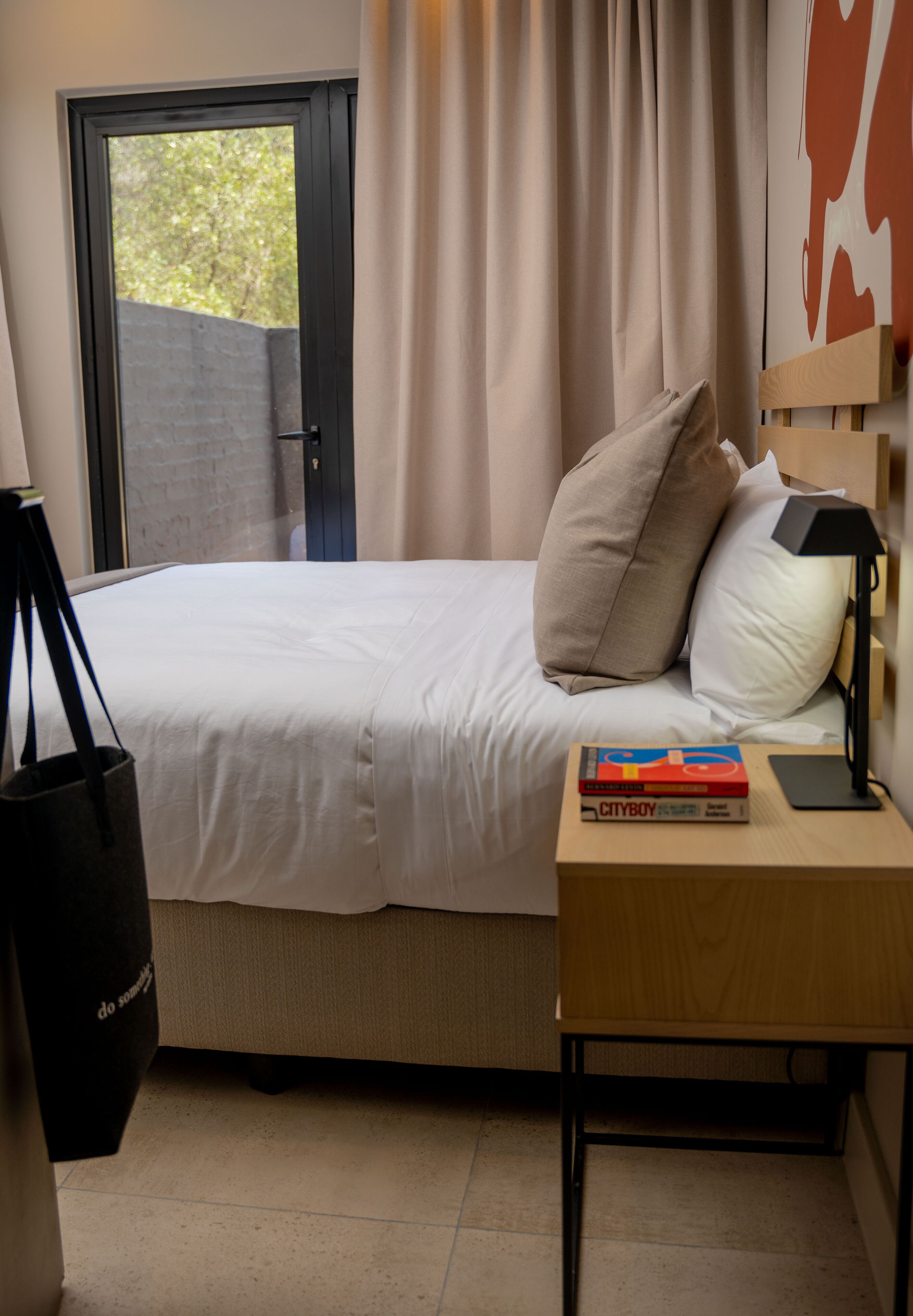 A neatly made bed with a white comforter and two pillows, a bedside table with a lamp and a book, and a door leading to an outdoor area.