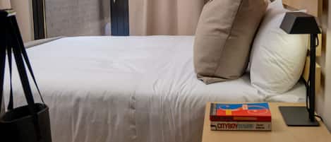 A neatly made bed with a white comforter and two pillows, a bedside table with a lamp and a book, and a door leading to an outdoor area.