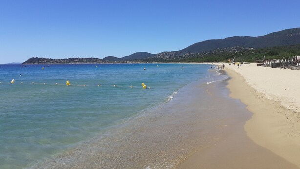 Une plage à l’eau d’un bleu limpide, bordée d’un rivage sablonneux, avec des collines en toile de fond.