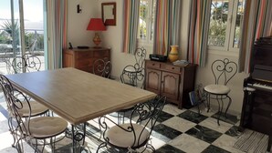 Un espace repas comprenant une table en bois, des chaises en métal, un piano et une vue sur la mer grâce à de grandes fenêtres.