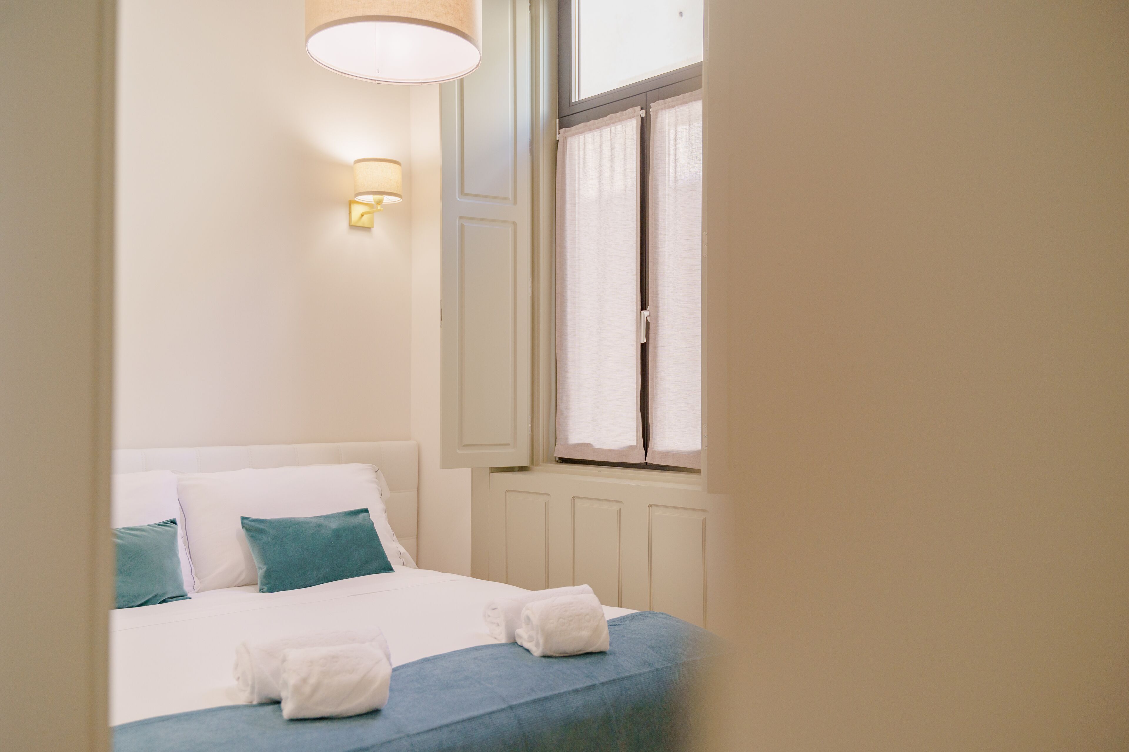 A neatly made bed with white and teal bedding, a wall-mounted light, and a window with curtains.