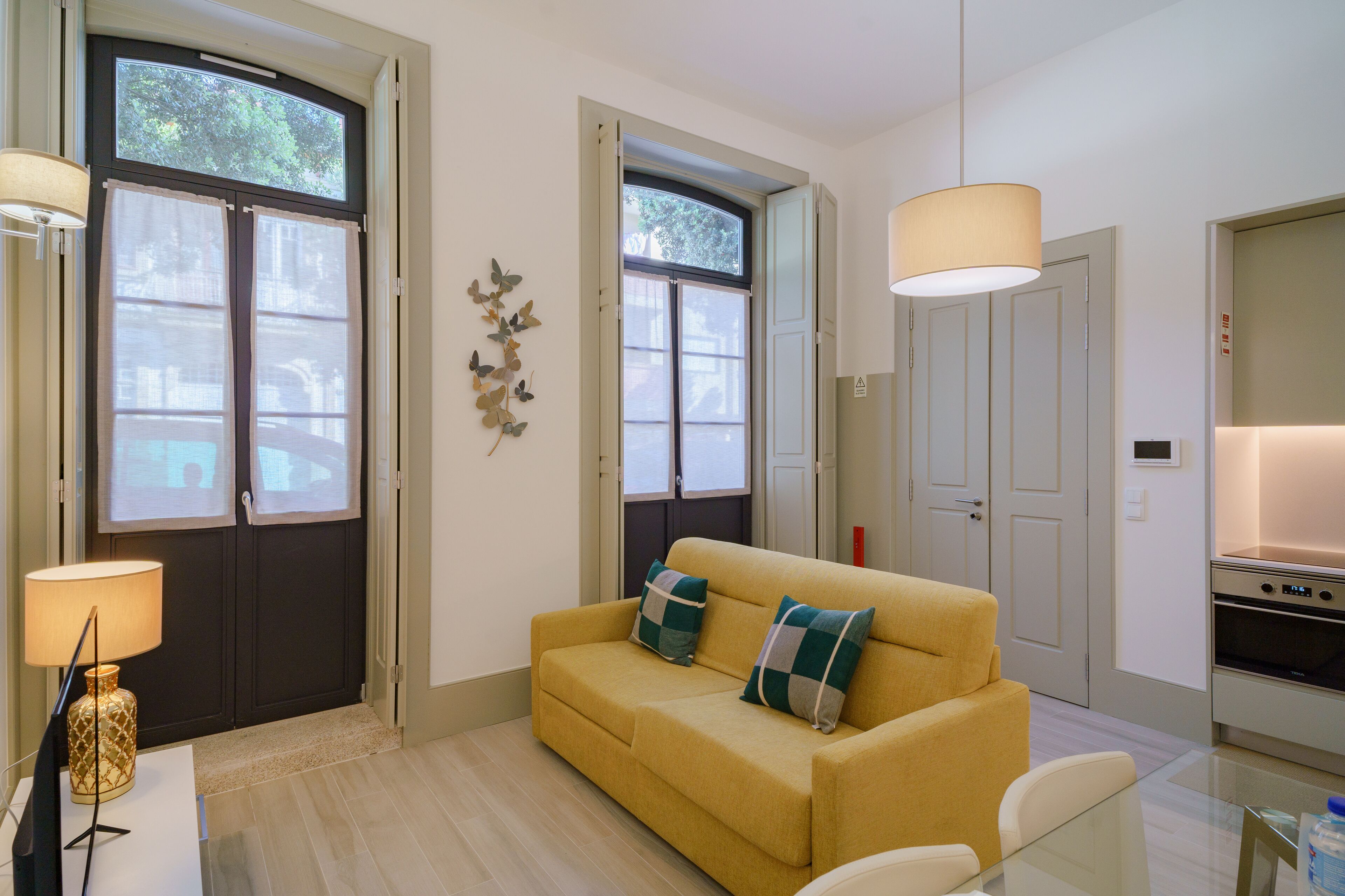 A modern living room with a sofa, a glass table, and a lamp. There are two windows with curtains, a door, and a kitchen area visible.