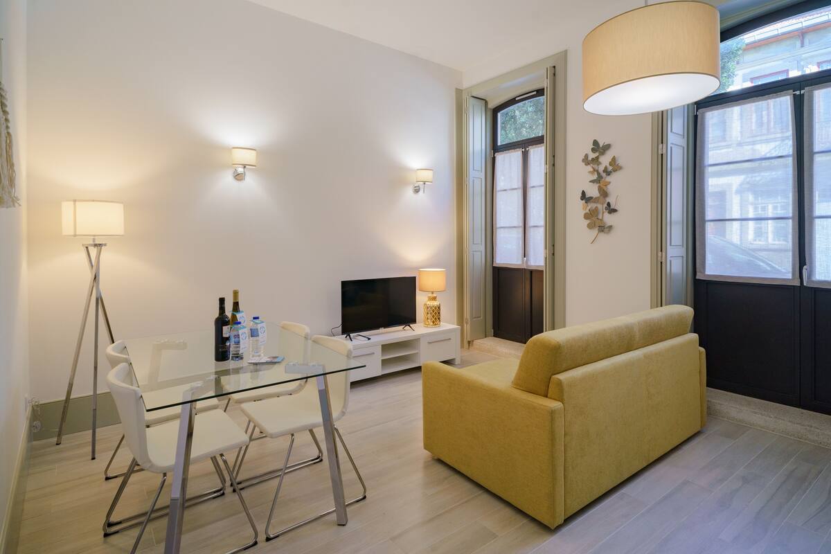 Una sala de estar moderna con una mesa de comedor de vidrio, sillas blancas, un sofá amarillo, un televisor de pantalla plana y un ventanal con vistas a edificios.