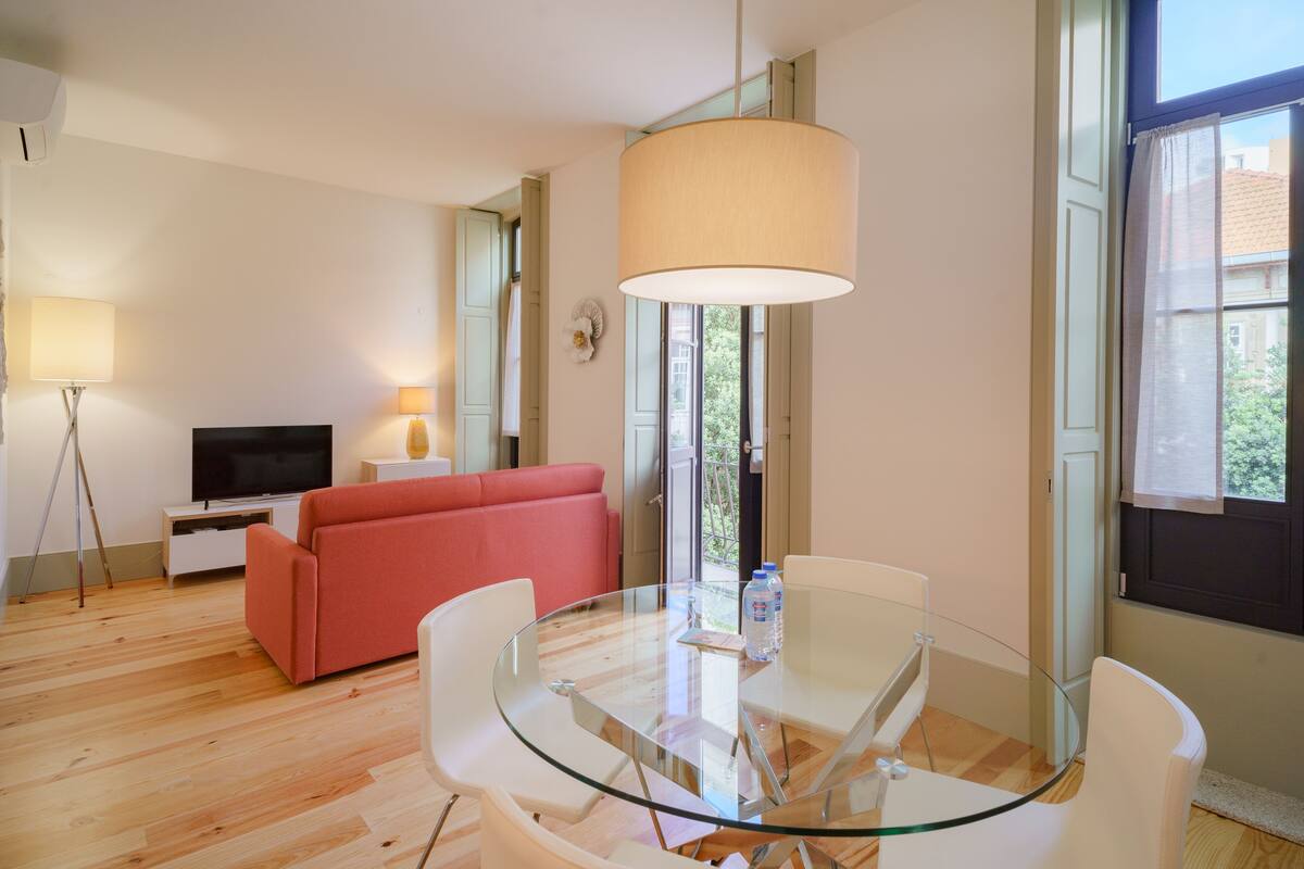 Una sala de estar moderna con una mesa de comedor de vidrio, un sofá rojo, un televisor de pantalla plana y un ventanal con cortinas.