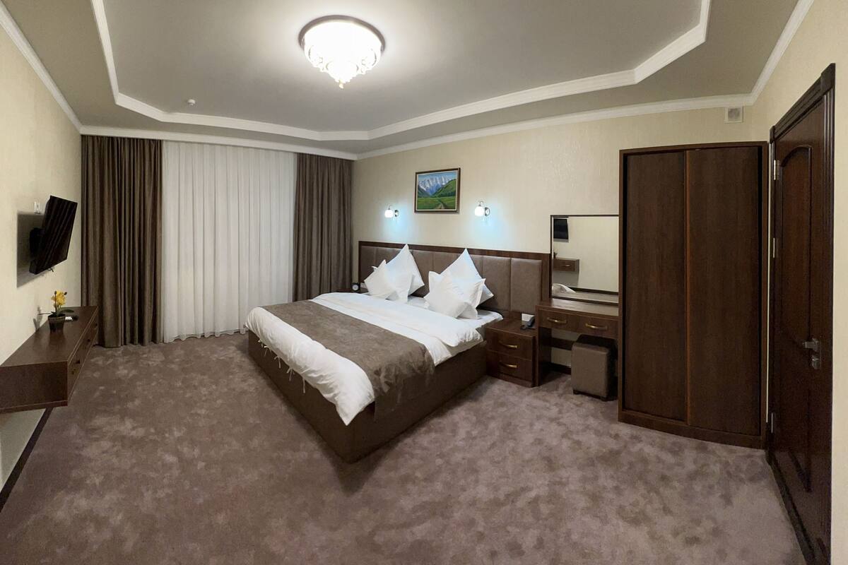 A hotel room with a large bed, a desk, a chair, a TV, and a wardrobe.