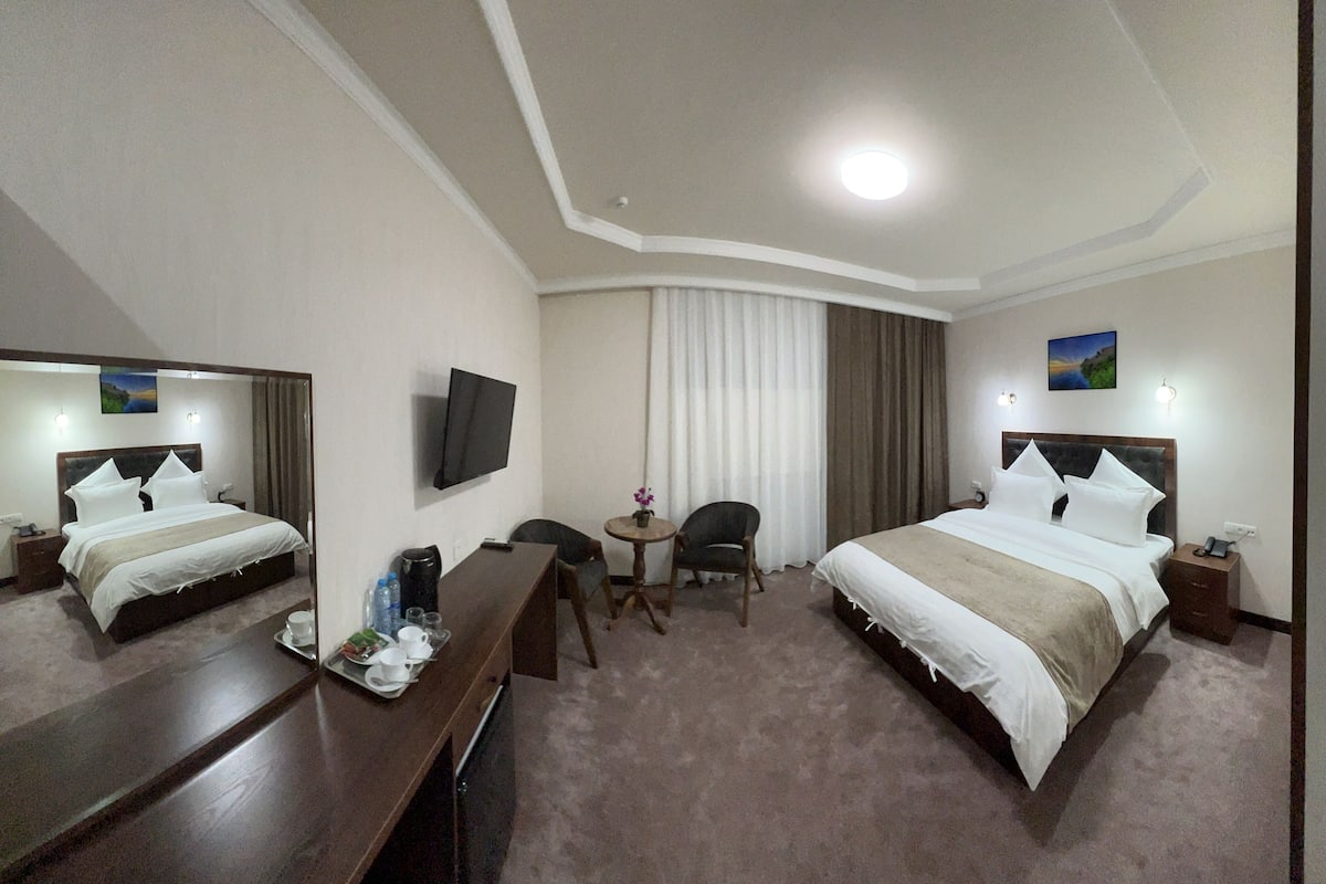 A hotel room with two beds, a desk, a chair, a TV, and a small table with a vase.