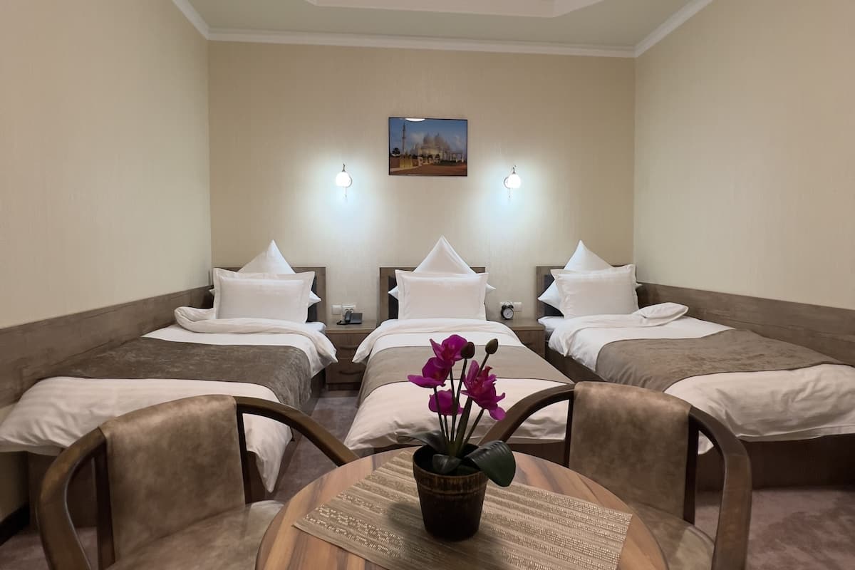 A hotel room with a round wooden table, two armchairs, a bed with white linens, and a small potted plant with purple flowers.