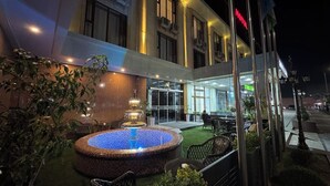 Ein Hotelgebäude bei Nacht mit beleuchtetem Springbrunnen, Außenbestuhlung und freiem Blick auf den Eingang.