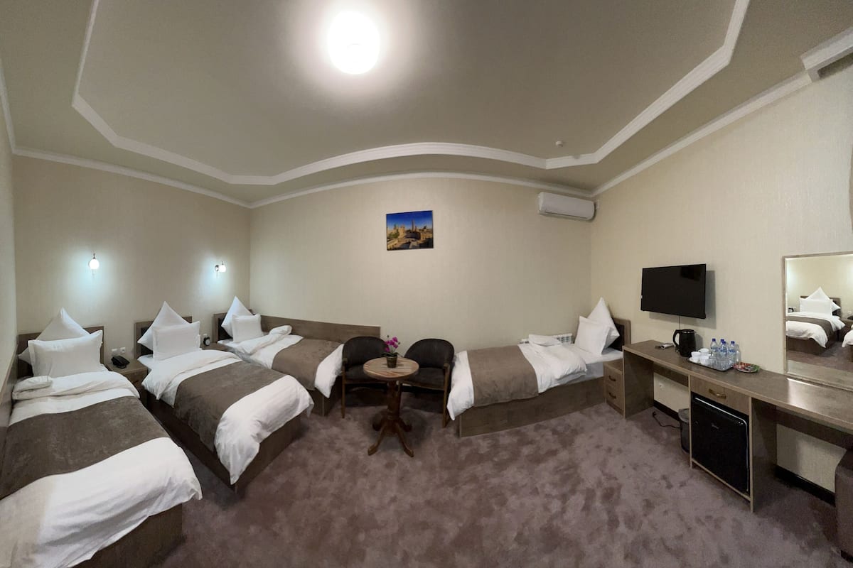 A hotel room with three single beds, a desk with a television, a small table with a vase, and a wall-mounted air conditioner.