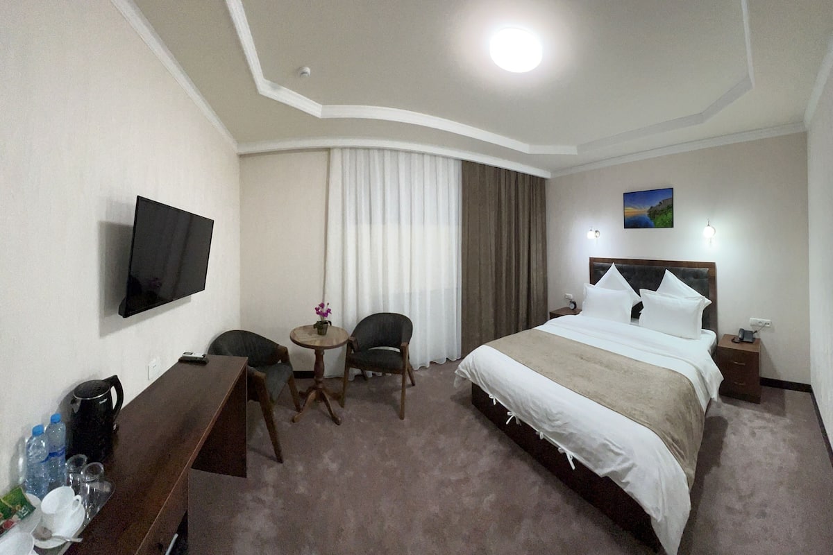 A hotel room with a bed, a TV, a desk, a chair, a small table with a vase, and a window with curtains.