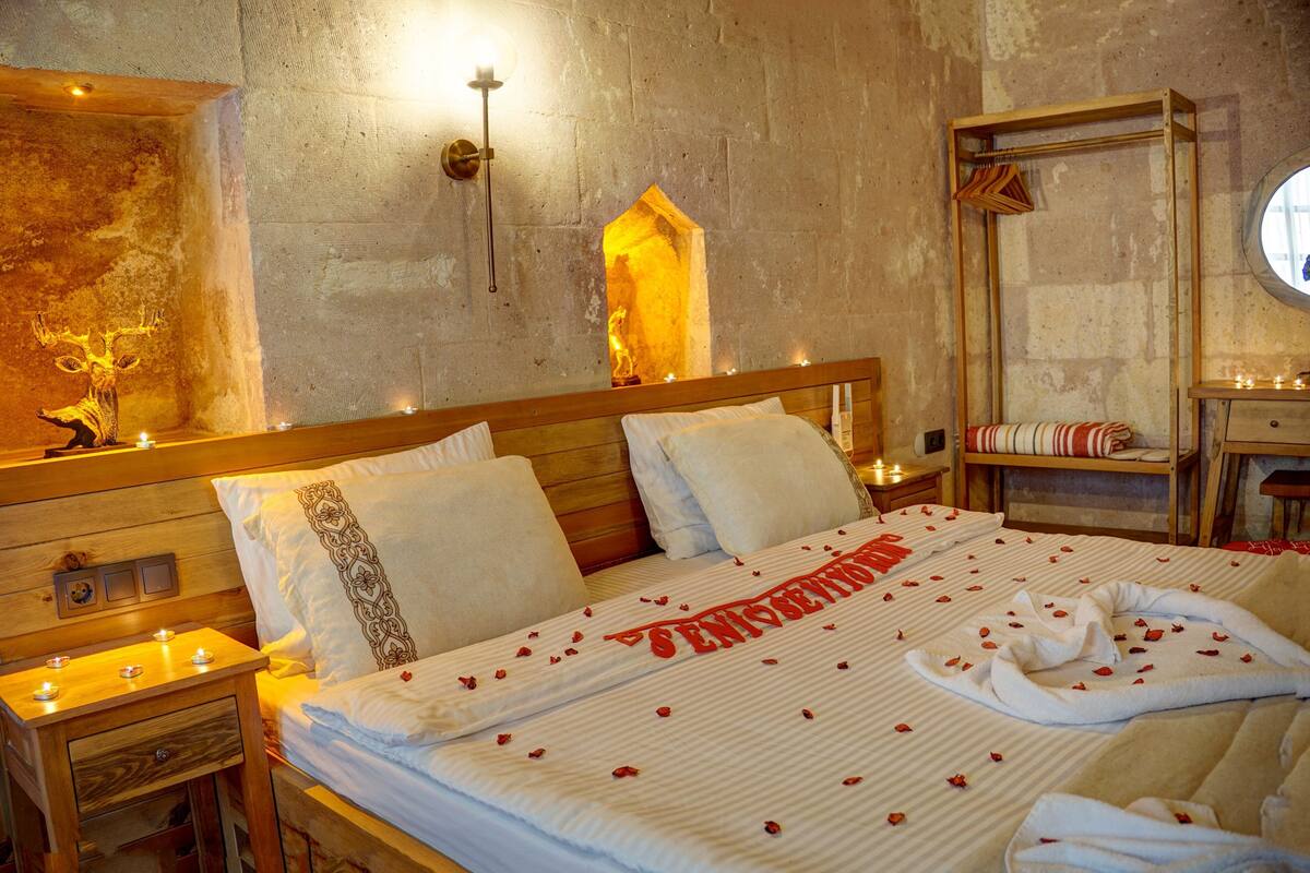A hotel room with a wooden headboard, a bed with rose petals and a towel, a nightstand with candles, and a stone wall with a small window.