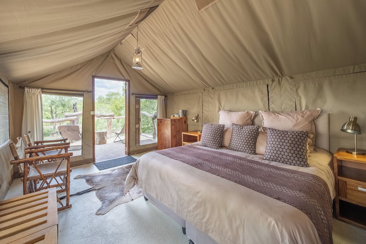 A tented room with a bed, wooden chairs, a desk, and a view of the outdoors.