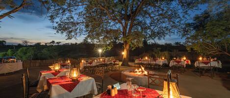 An outdoor dining area with tables set for a meal, surrounded by trees and a clear sky.