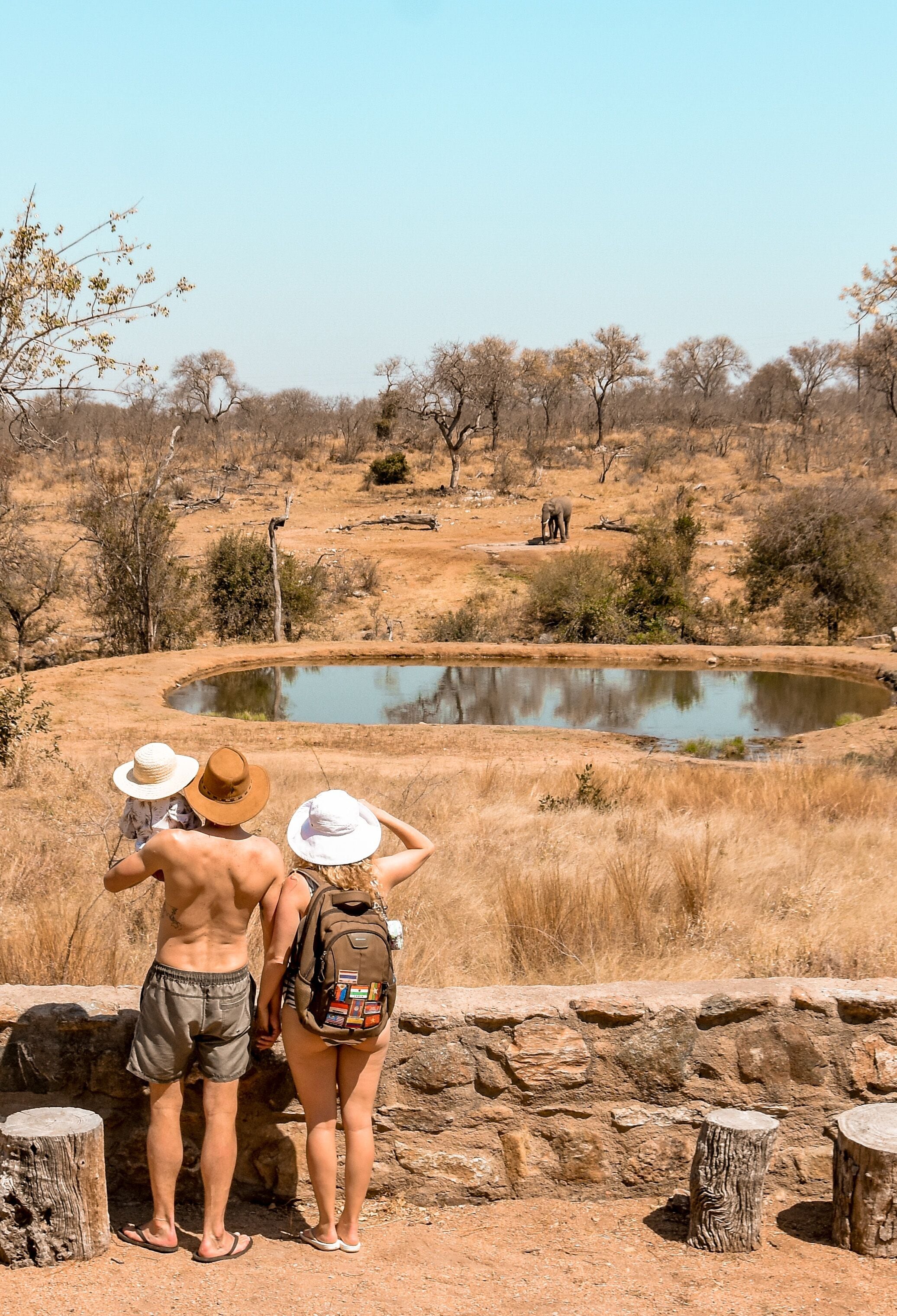 A couple standing by a pond with a stone wall and wooden posts in a dry, open landscape.