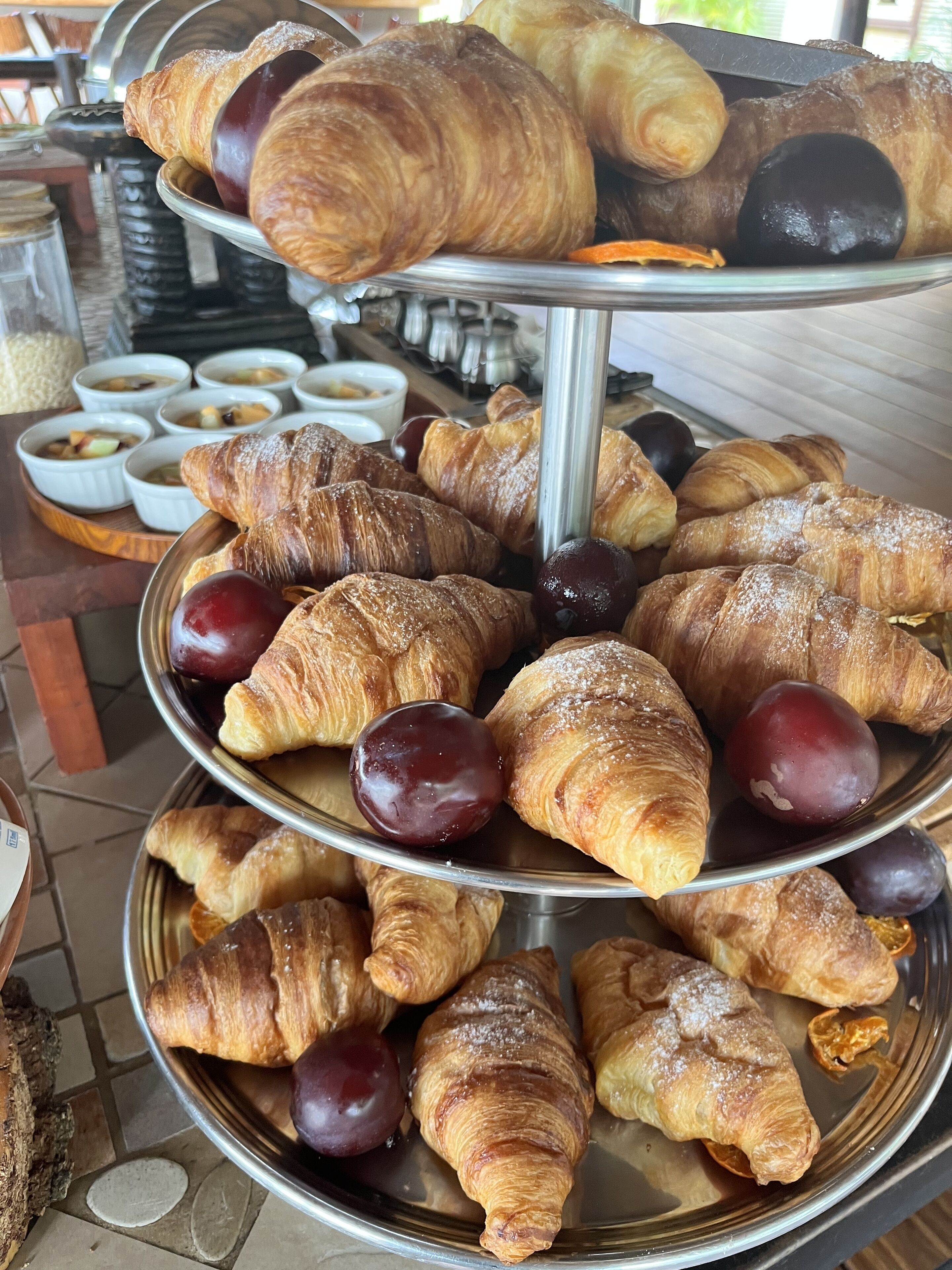 A multi-tiered display of pastries and fruits, including croissants and plums, on a metal stand.