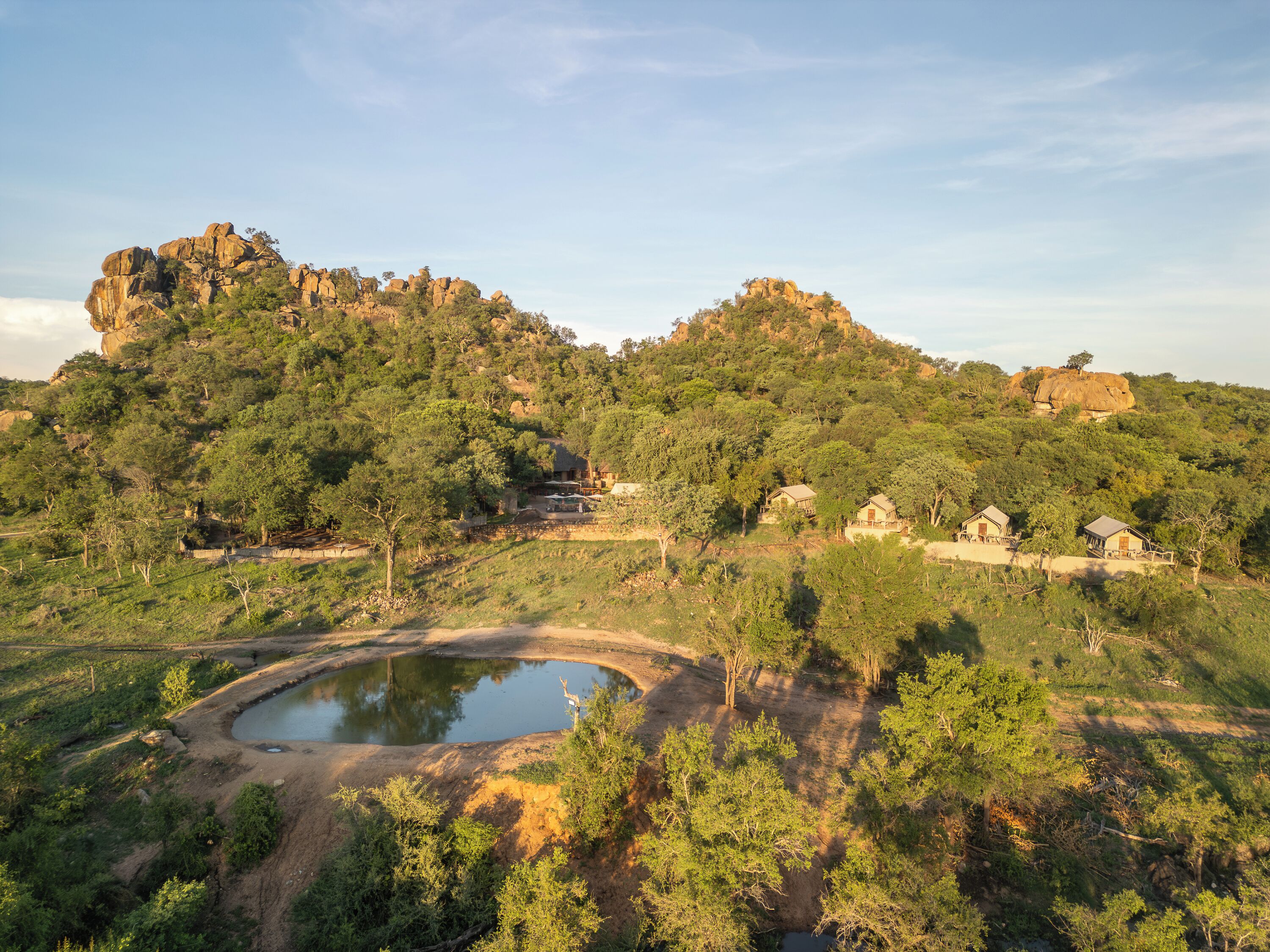 A natural landscape with a pond, rocky hills, and several buildings nestled among trees.
