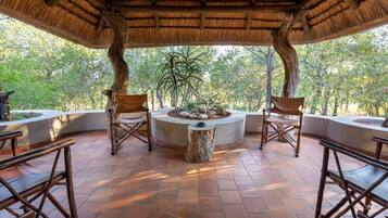 Relax in a shaded seating area surrounded by lush trees and foliage, with a thatched roof and comfortable seating for two.