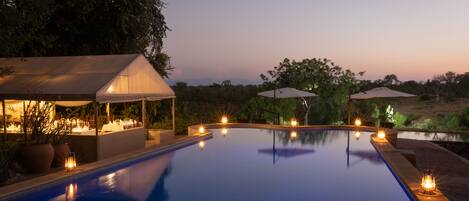 A swimming pool with a covered seating area and umbrellas, surrounded by trees and a view of the landscape.