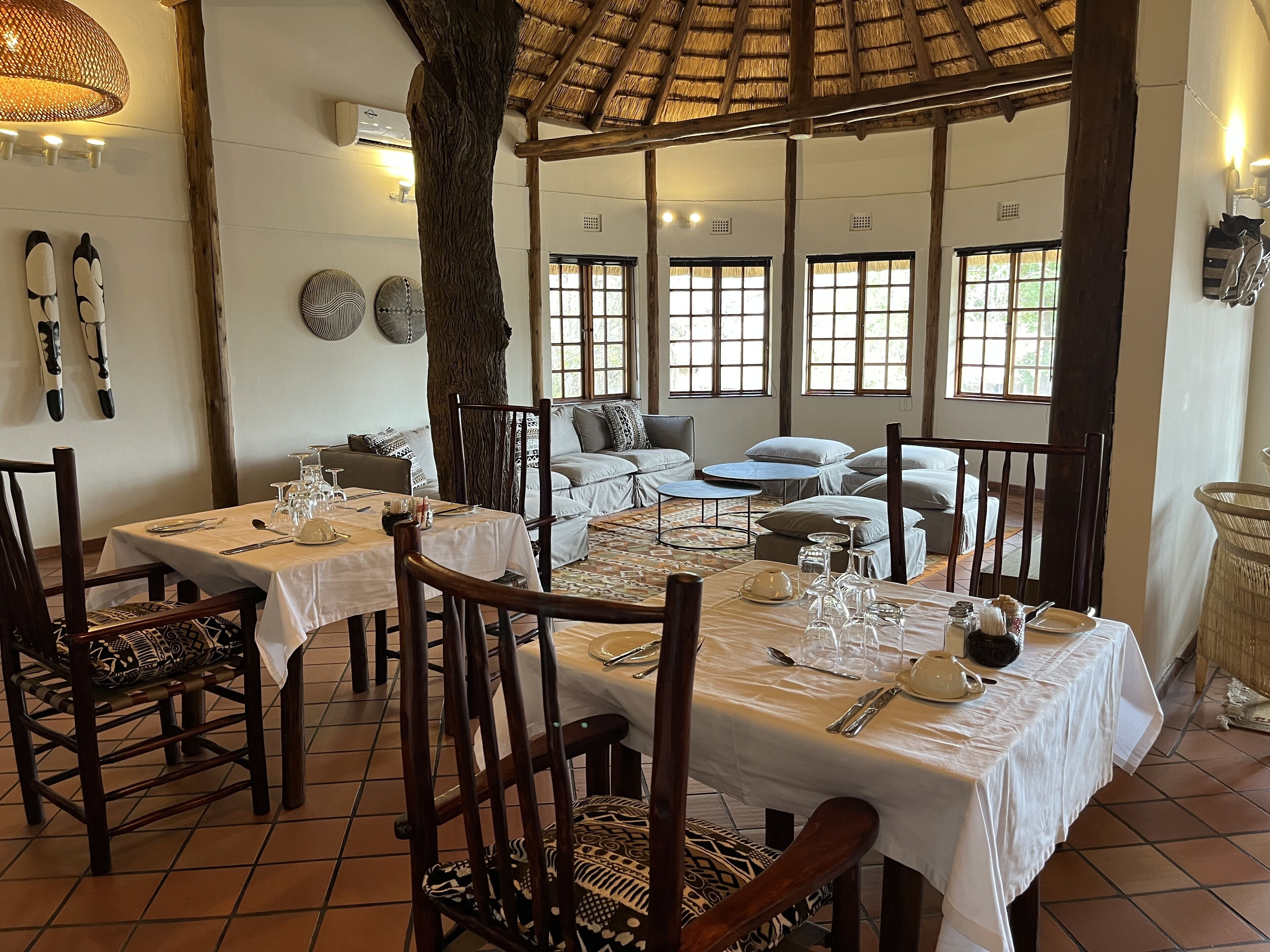 A dining area with tables set for a meal, a large tree trunk in the center, and a view of the outdoors through large windows.