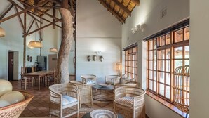 Una habitación amplia con techo alto, vigas de techo tejidas y un ventanal con vistas. Hay sillas de mimbre y una mesa de comedor con un cuenco de fruta.