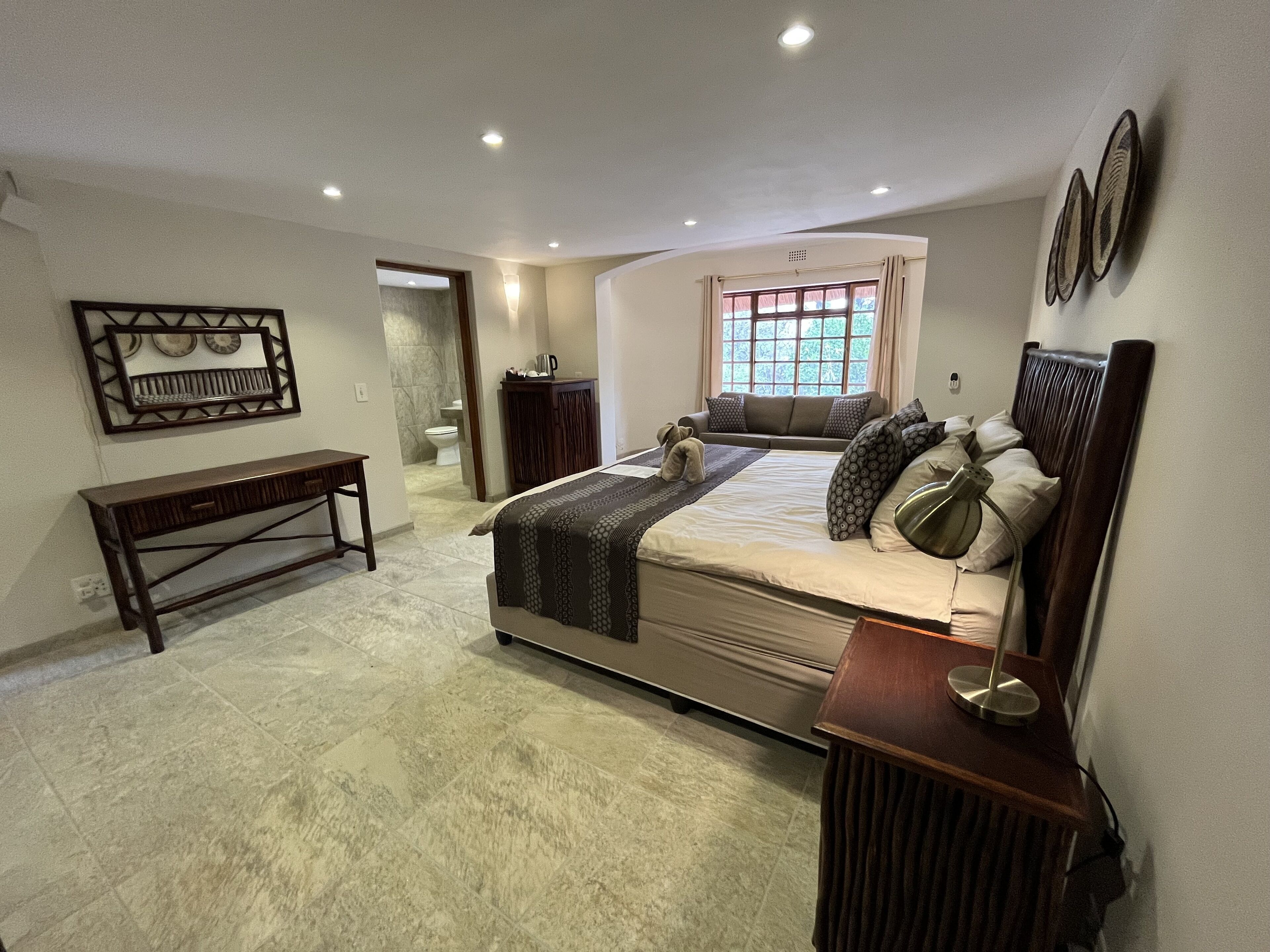 A bedroom with a large bed, bedside tables, a bench, a window with curtains, and a bathroom visible in the background.