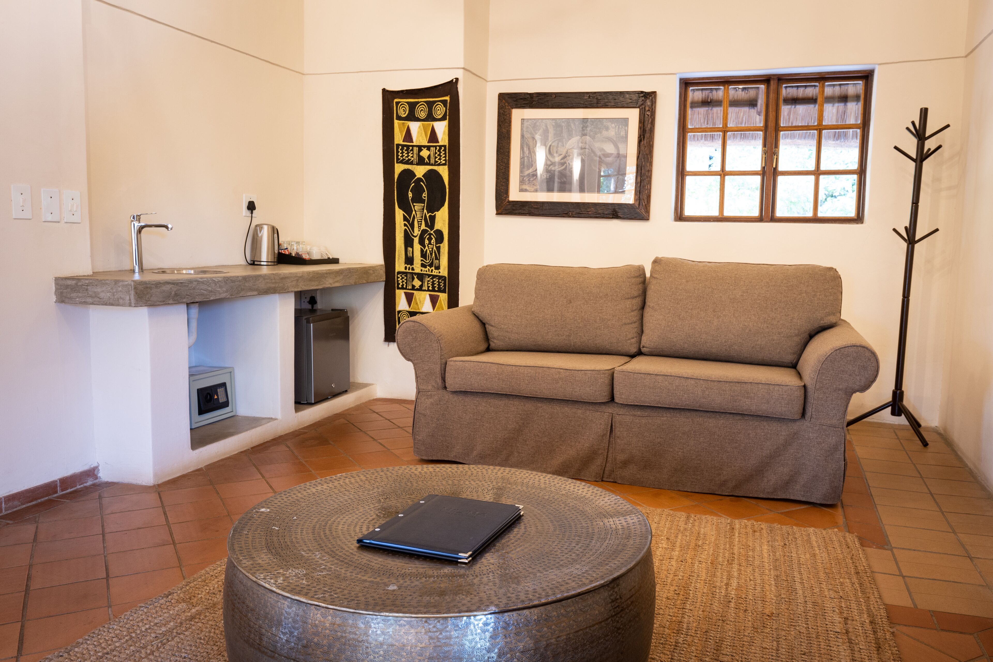 Una sala de estar con un sofá, una mesa de centro, una pequeña cocina con un lavamanos y un hervidor, un cuadro en la pared y una ventana con vistas.