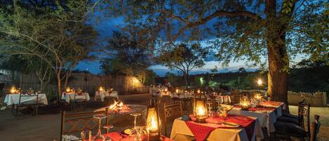 An outdoor dining area with tables set for a meal, surrounded by trees and a wooden fence.