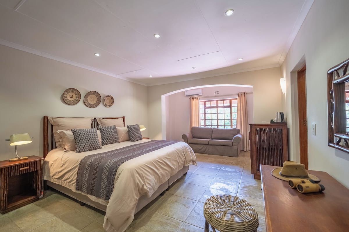 A bedroom with a bed, bedside tables, a sofa, a wicker stool, and a desk with a hat.