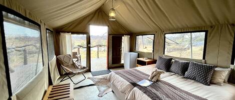 A tented accommodation room with a bed, a chair, a desk, and a view of the outdoors.