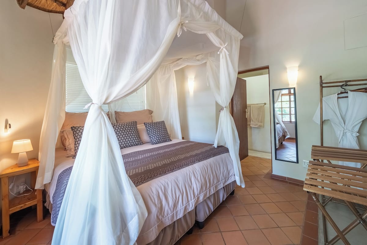 A bedroom with a four-poster bed, a nightstand with a lamp, a wooden bench, and a mirror on the wall.