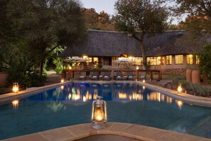 A swimming pool with a lantern in the foreground, surrounded by lounge chairs and a building with a thatched roof in the background. - MILIMA Big 5 Safari Lodge (Hoedspruit)