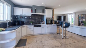 A modern kitchen with white cabinets, a black backsplash, and a central island. There is a dining area with a bar stool and a bookshelf in the background. - VILLA L'ESTEL in the countryside with private pool 10 minutes from Arles (Tarascon)