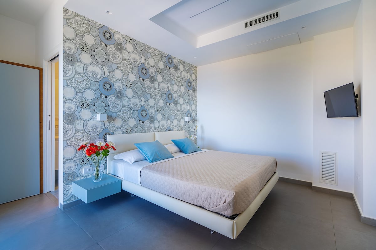 Ein modernes Schlafzimmer mit ornamentalem Wandpapier, einem Bett mit blauen Kissen, einem Nachttisch mit Blumen und einem an der Wand montierten Fernseher.