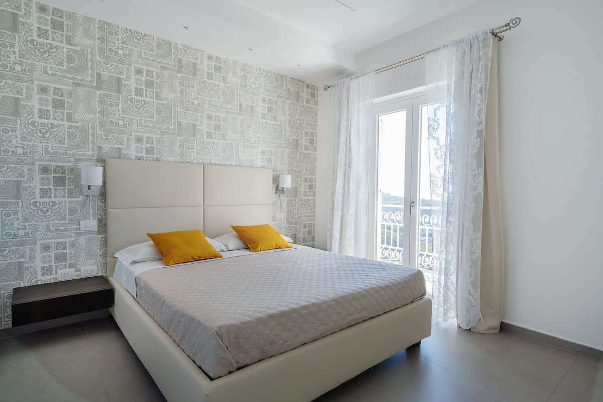 Ein Schlafzimmer mit gemusterten Wänden, einem Bett mit weißer Bettwäsche und zwei gelben Kissen, einem Nachttisch und einem Fenster mit Vorhängen.