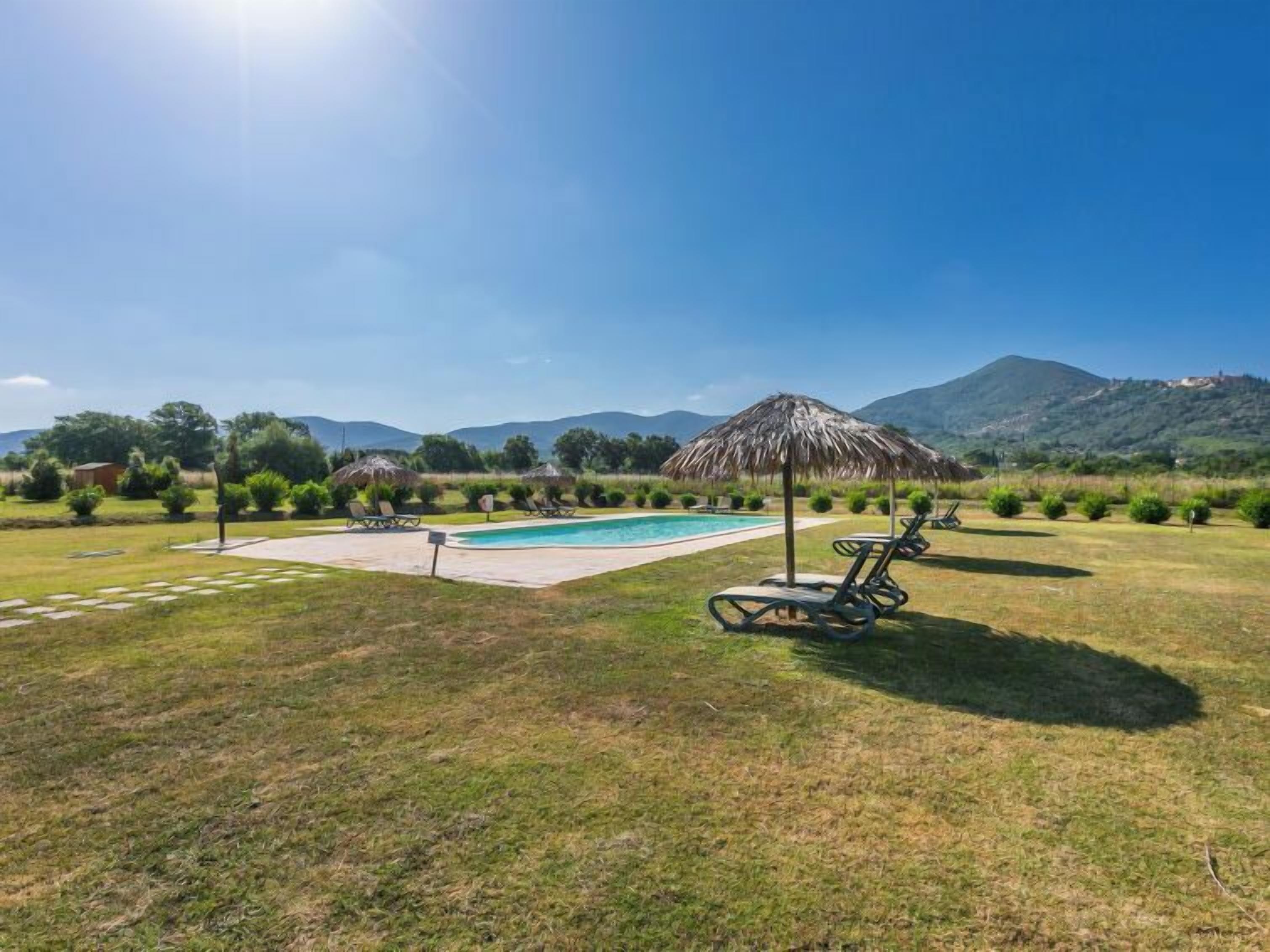 A swimming pool with lounge chairs and a thatched umbrella, surrounded by grass and mountains in the background.