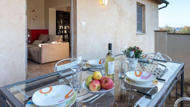 Un espacio de comedor al aire libre con mesa de vidrio, platos, cubiertos, una botella de vino y un cuenco de fruta.