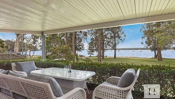 Un espace extérieur aménagé avec des meubles en osier, une table à plateau de verre et une vue sur un lac où naviguent des bateaux.