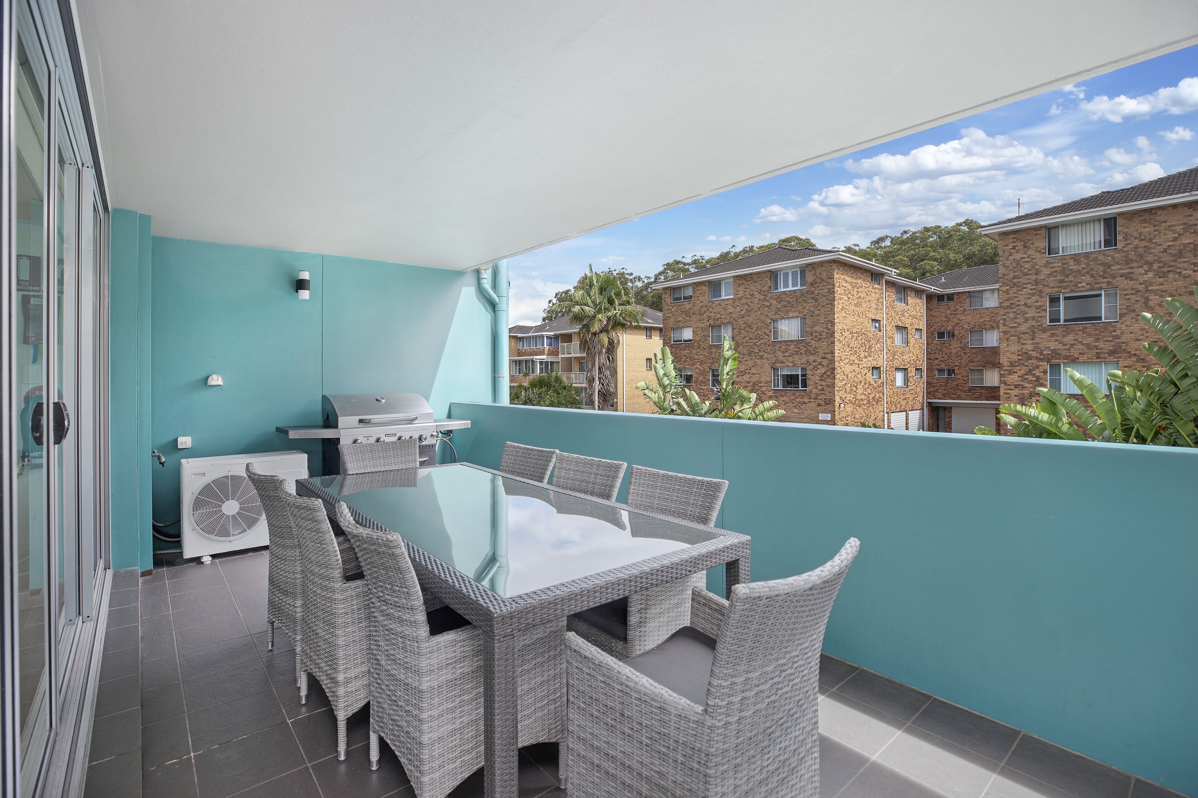 Un balcon avec une table à plateau en verre et des chaises en osier, un barbecue et un appareil de climatisation.
