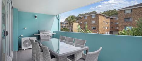 Un balcon avec une table à plateau en verre et des chaises en osier, un barbecue et un appareil de climatisation.