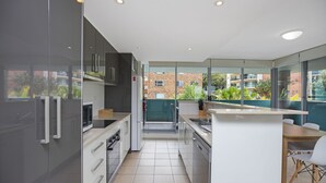 A modern kitchen with a central island, white cabinetry, and a view of a brick building through large windows. - Spacious. Coastal. Fresh. (Forster)